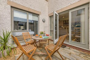 Eine Terrasse mit einem runden Tisch, vier Holzstühlen, einem Blumentopf und einer Glastür, die zu einem Innenraum führt.
