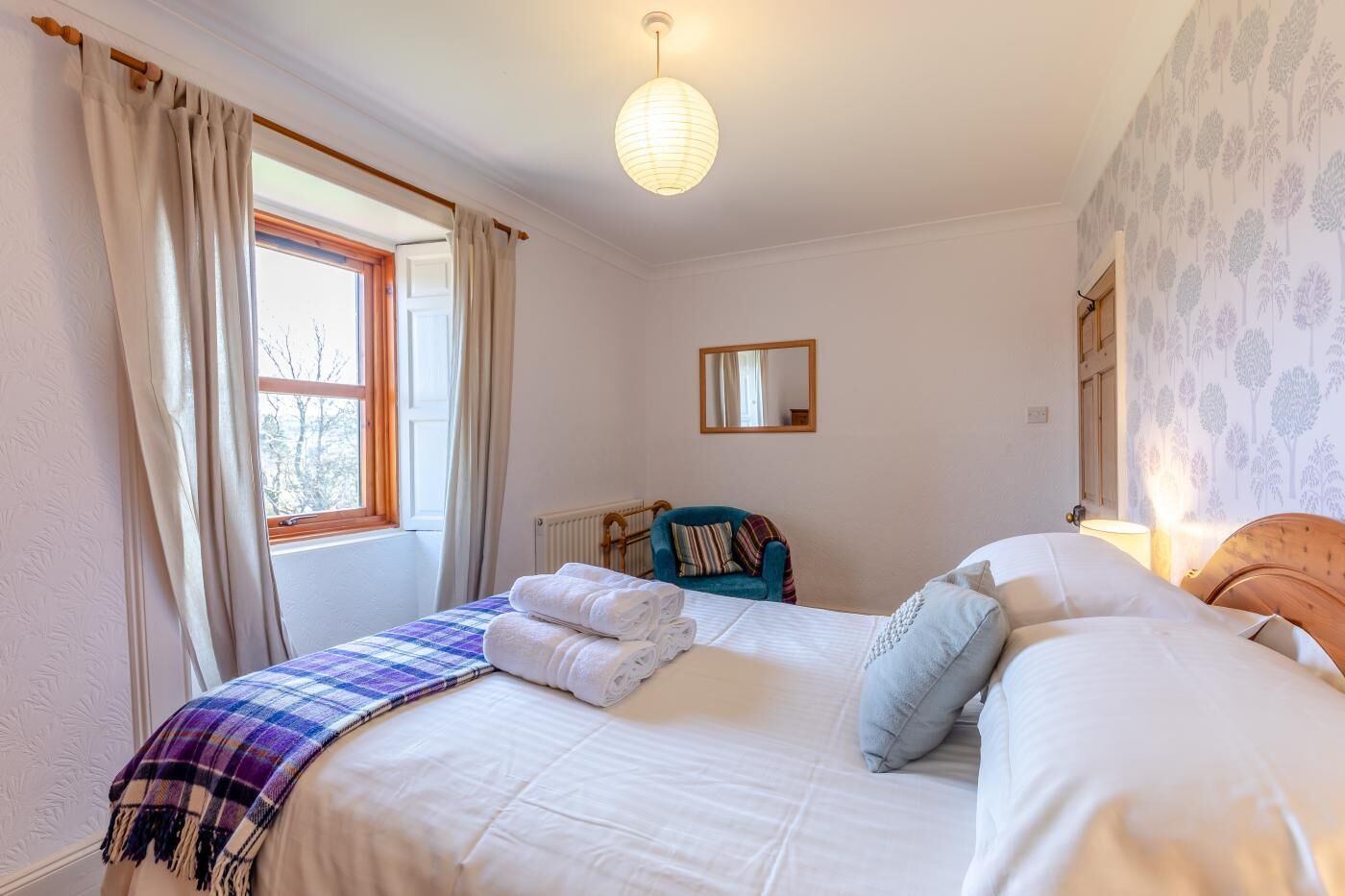 A bedroom with a bed, a window with curtains, a chair, a radiator, and a wall-mounted mirror.