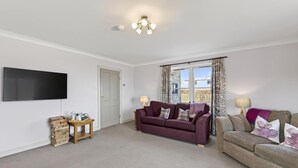 A living room with a flat-screen TV, two sofas, a wooden side table, and a window with curtains. - 5 Bed in Cockburnspath (oc-r35051) (Cockburnspath)