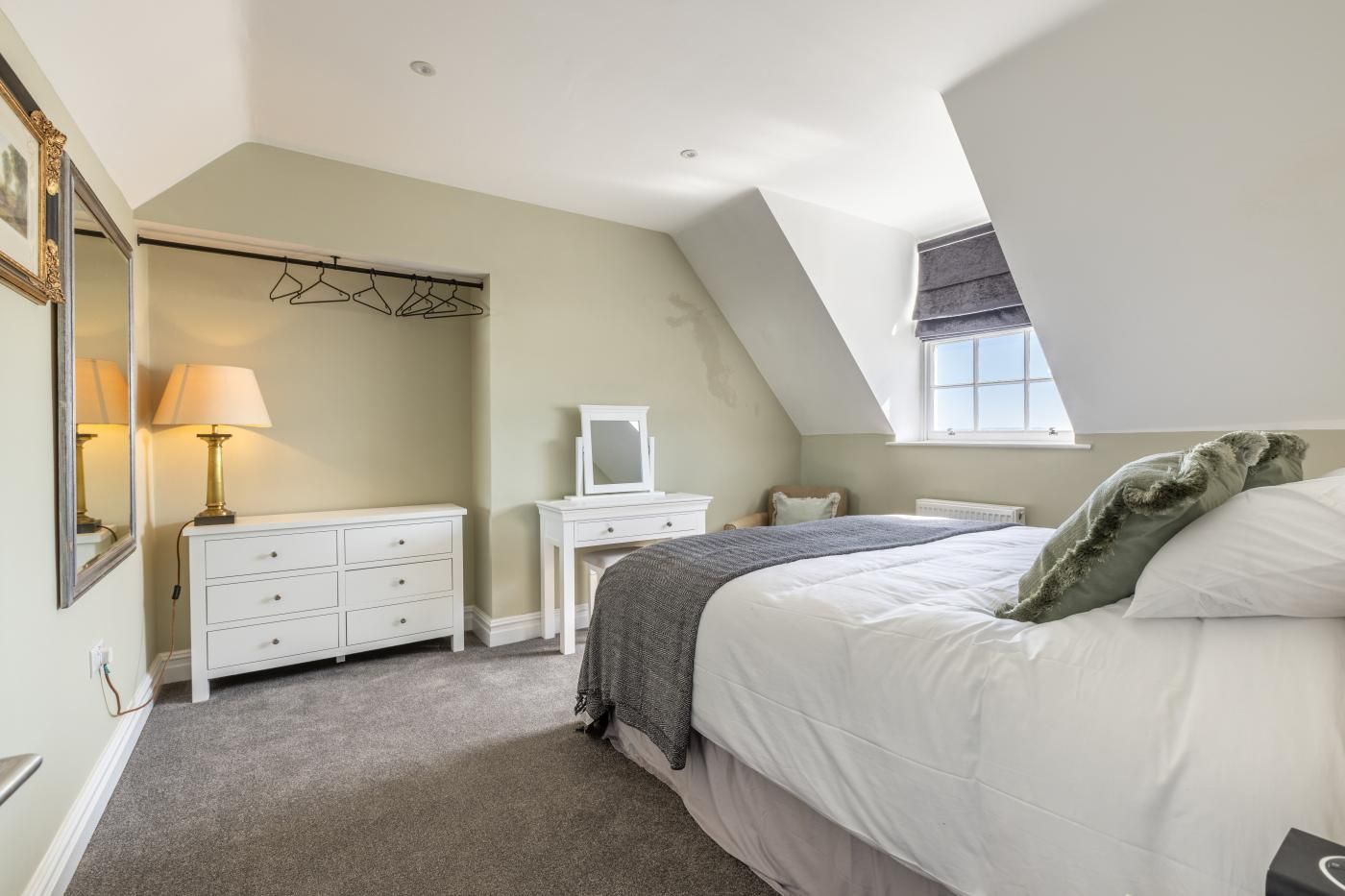 Une chambre avec un plafond en pente, un lit avec une literie blanche, une commode, un miroir et une fenêtre.