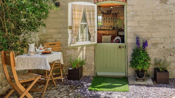 Un edificio rústico de piedra con una puerta verde, una pequeña mesa con mantel blanco y plantas en macetas.