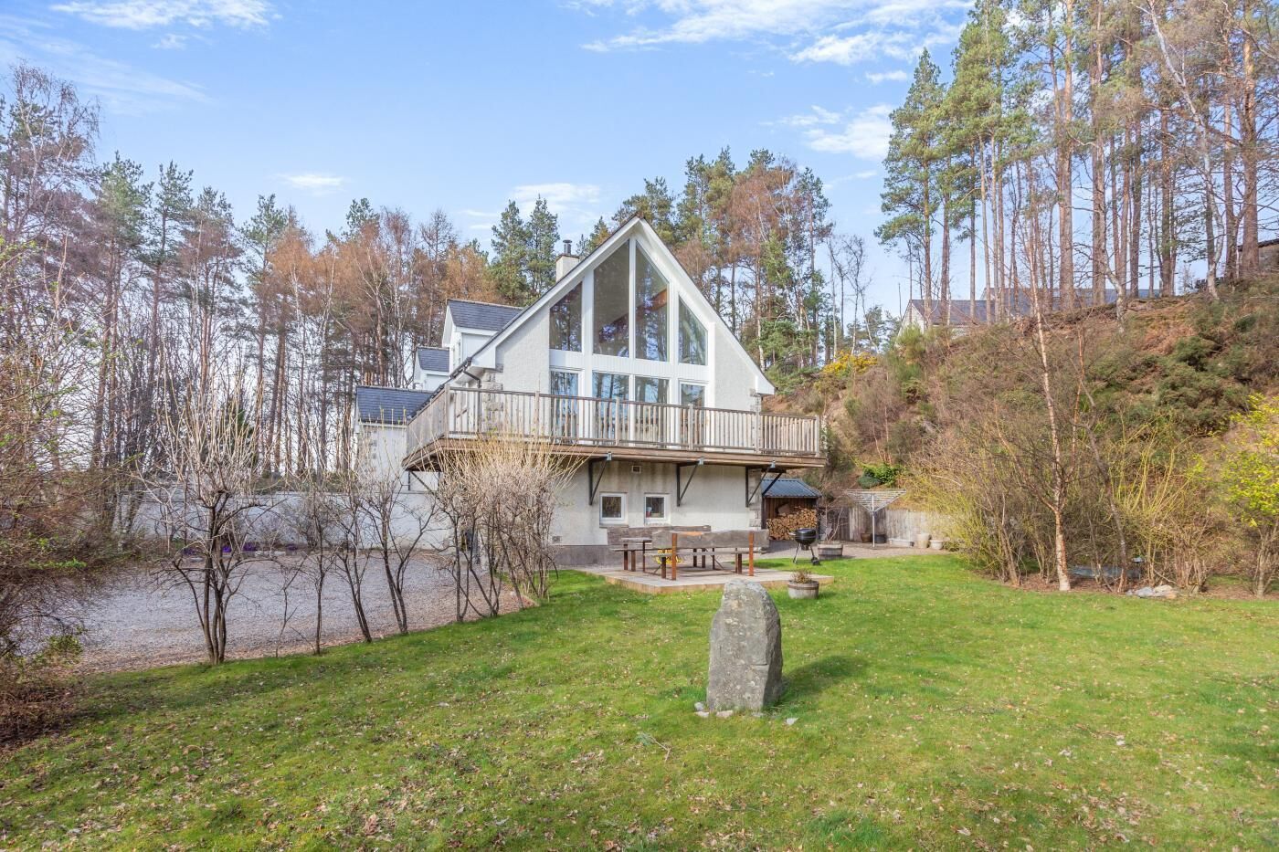 Une maison moderne dotée d’une grande terrasse, avec une pierre au premier plan et une zone boisée en arrière-plan.