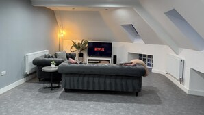 Ein modernes Loft-Zimmer mit einem großen grauen Ecksofa, einem kleinen Beistelltisch, einer Stehlampe und einem Flachbildfernseher, der 'NETFLIX' anzeigt.