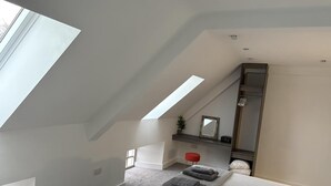 Ein Schlafzimmer mit schrägen Wänden, einem Oberlicht, einem Bett mit grauer Bettwäsche, einem roten Hocker, einer kleinen Pflanze und einem Spiegel.