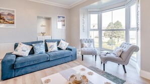 Ein Wohnzimmer mit blauem Sofa, grauen Sesseln, einem weißen Couchtisch und einem Erkerfenster mit Blick.