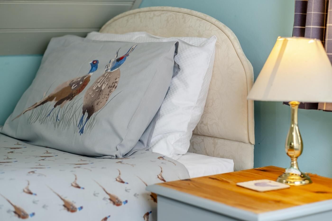Ein Bett mit gemusterten Textilien, ein Kissen mit Vogelmotiven, eine Nachttischlampe und ein Nachttisch mit einem kleinen Gegenstand darauf.