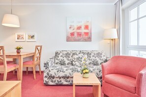 A living room with a sofa, a red armchair, a wooden table, and a dining area with chairs and a table. - Ferienwohnung Sellin Nahe Strand mit Balkon (Sellin)