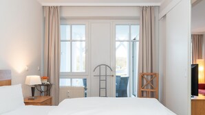 A hotel room with a bed, a nightstand with a lamp, a chair, a TV, and a view of the outdoors through large windows. - Strandurlaub mit der Familie - Ferienwohnung mit Balkon in Sellin (Sellin)