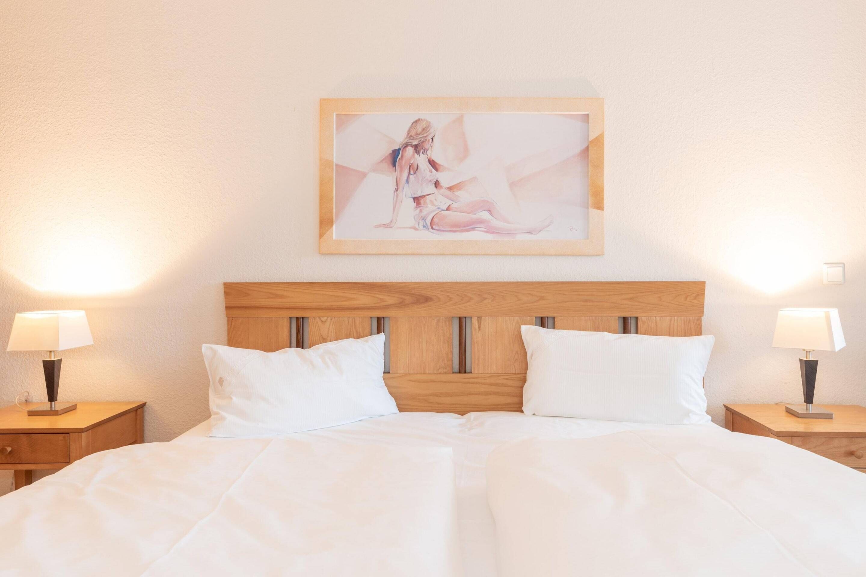 Ein Hotelzimmer mit hölzernem Kopfteil, weißer Bettwäsche, zwei Nachttischlampen und einem gerahmten Kunstwerk an der Wand.