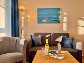 A living room with a sofa, a coffee table with a bottle and glasses, a painting of seagulls, and a lamp. - Seepark Sellin Fewo fur 3 Personen (Sellin)