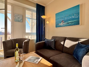 A living room with a sofa, a table with a bottle and glasses, a painting of seagulls, and a window with curtains. - Seepark Sellin Fewo fur 3 Personen (Sellin)