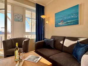 Ein Wohnzimmer mit einer Couch, einem Tisch mit Flasche und Gläsern, einem Gemälde von Möwen und einem Fenster mit Vorhängen.