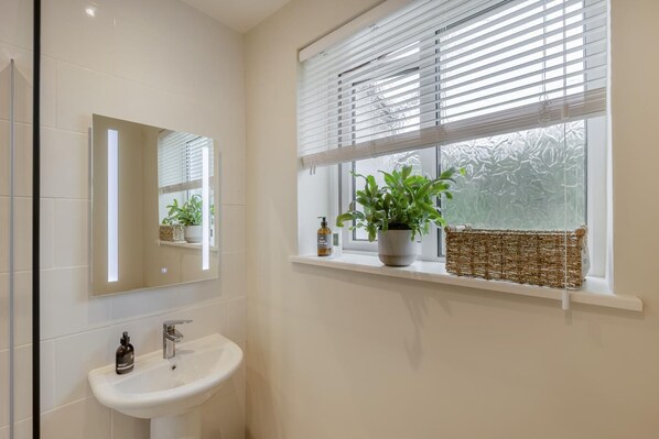 A bathroom with a white sink, a mirror, a window with blinds, and a shelf with a plant and a bottle.