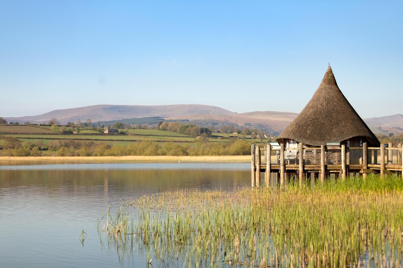 A thatched-roof structure by a calm lake with a wooden deck, surrounded by grassy fields and distant hills.