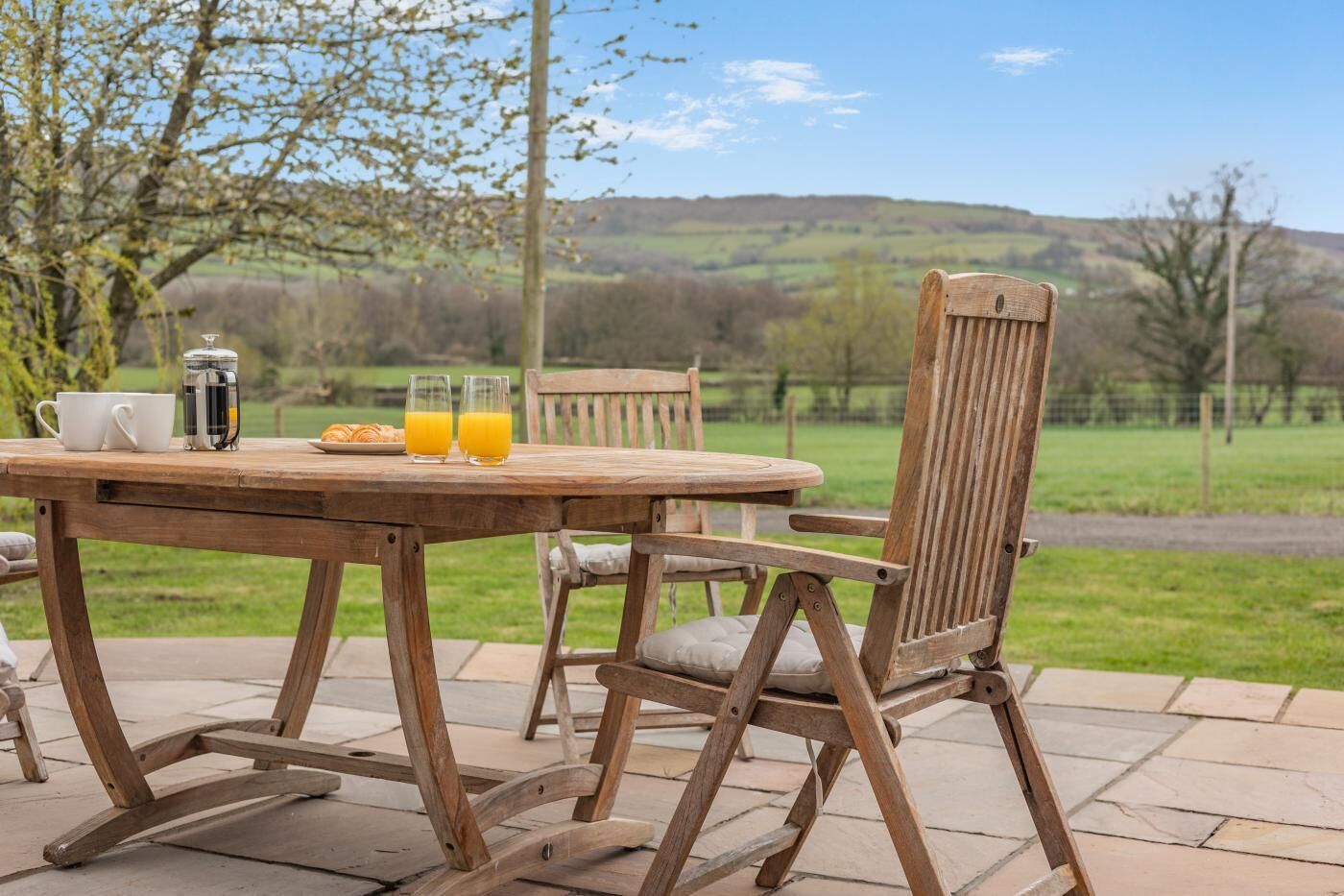 Una mesa de madera al aire libre con sillas, una cafetera y vasos de jugo de naranja.