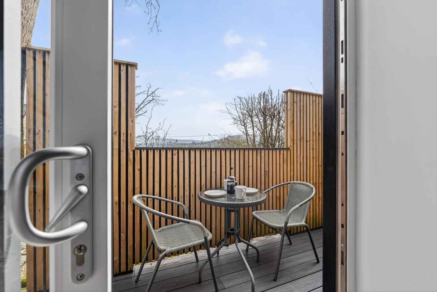 Een houten terras met twee rieten stoelen en een kleine tafel, een metalen deur, en uitzicht op een houten schutting en bomen.