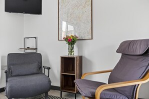 Ein Zimmer mit grauem Sofa, einem Holzstuhl, einer kleinen Holzregal mit Blumenvase, einer Karte an der Wand und einem darüber montierten Fernseher.