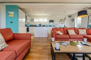 A modern living room with a sofa, a coffee table with magazines and fruit, and a kitchen in the background. - 3 Bed in Portreath (oc-b38226) (Portreath)