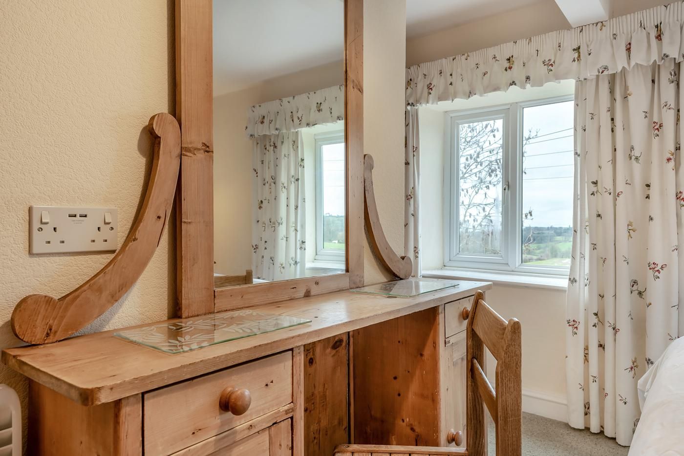 A wooden dresser with a mirror, a window with floral curtains, and a view of the outdoors.