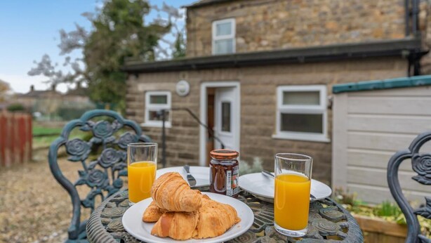 Desayuno con cruasanes, zumo de naranja y un tarro de mermelada sobre una mesa de metal frente a una casa de piedra.
