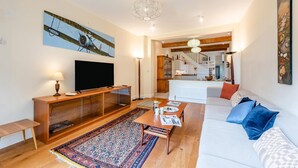 Una sala de estar con televisor, muebles de madera, alfombra estampada y una escalera que lleva a una cocina.