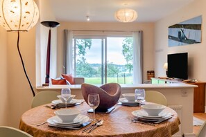 A dining area with a table set for four, a large window with a view of greenery, a television on a stand, and a modern pendant light.