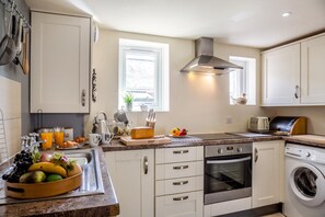 A modern kitchen with white cabinets, a stainless steel range hood, a built-in oven, a washing machine, and a breakfast setup with fruits and juice. - 1 Bed in Llandwrog (oc-h36065) (Llandwrog)