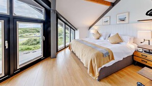 A bedroom with a large bed, wooden beams, a view through large windows, and a bedside table with a lamp. - 2 Bed in Borth (oc-ystiw4) (Glanwern)