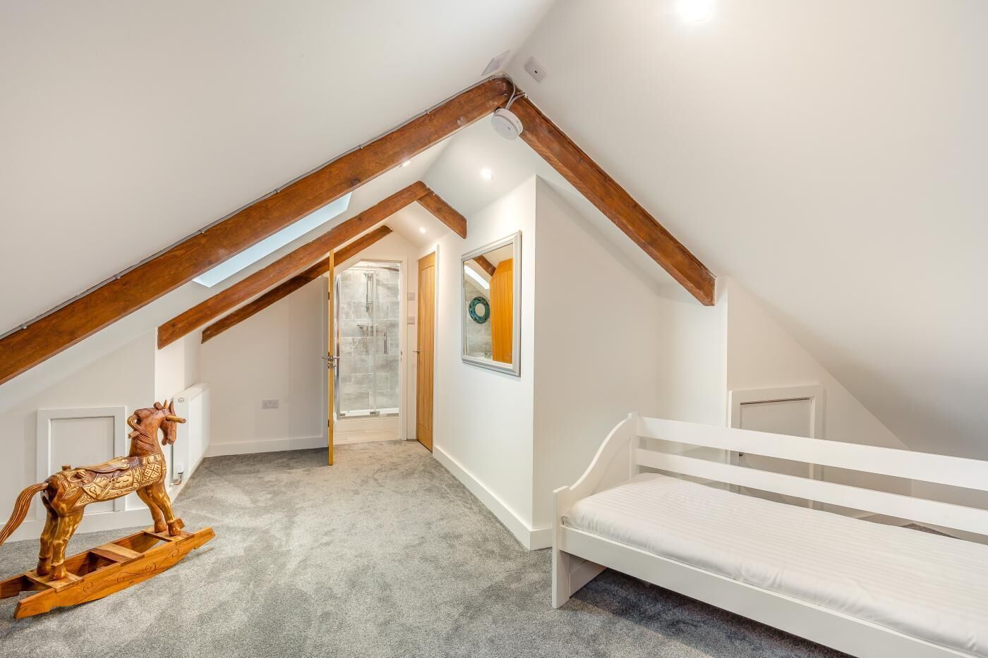 A bedroom with a sloped ceiling, a wooden rocking horse, a white bed, and a door leading to another room.