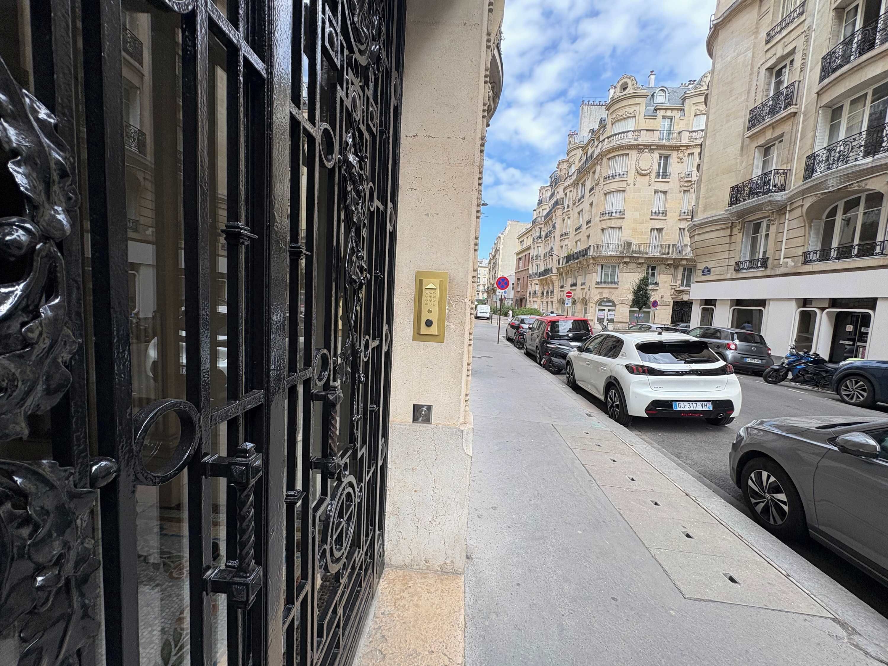 A street view with parked cars, a building with ornate black gates, and classic European architecture.