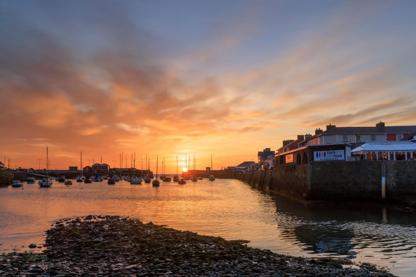 Un port au coucher du soleil, avec des bateaux, un mur de pierre et des bâtiments portant des enseignes.