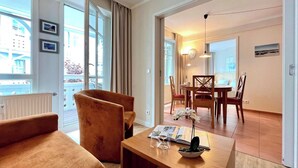 A modern hotel room with a sofa, armchair, dining table, and a view of the city through large windows. - Urlaub auf Rugen in Einer Ferienwohnung (Sellin)