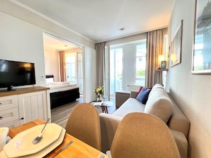A hotel room with a bed, a TV, a dining table with a plate and spoon, a sofa, and a window with curtains. - Hochwertige Ferienwohnung fur 3 im Seepark Sellin (Sellin)