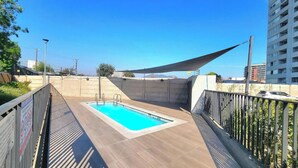 Una piscina en la azotea con cubierta de madera, rodeada por una barandilla de metal y una vela de sombra.