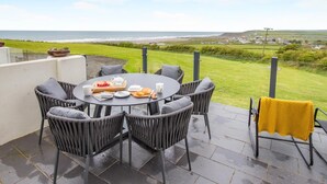 Une table ronde entourée de chaises, un plateau avec de la nourriture et des boissons, et une vue sur la mer et le paysage.