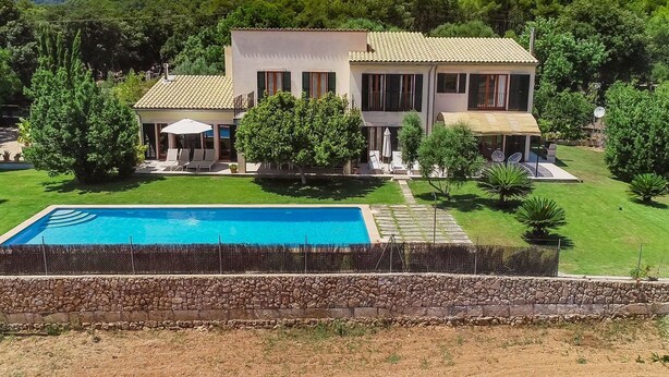 Una casa de dues plantes amb una piscina, envoltada de vegetació i turons.
