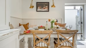 Un coin repas avec une table blanche, des chaises en bois et un banc. On y trouve deux lampes fixées au mur, un tableau encadré et un vase avec des fleurs sur la table.