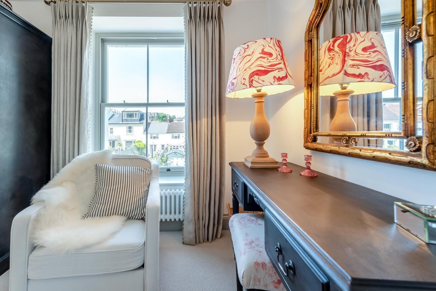 Ein Zimmer mit Blick auf Gebäude, ein weißer Stuhl mit gestreiftem Kissen, eine Kommode mit Spiegel und zwei Lampen mit Marmormuster.