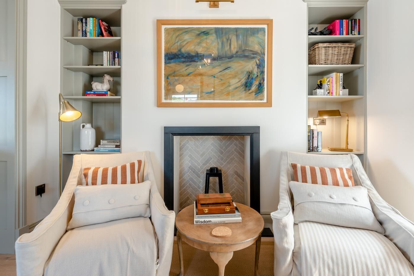 A room with two armchairs, a small table with books, a fireplace, and bookshelves with books and decorative items.