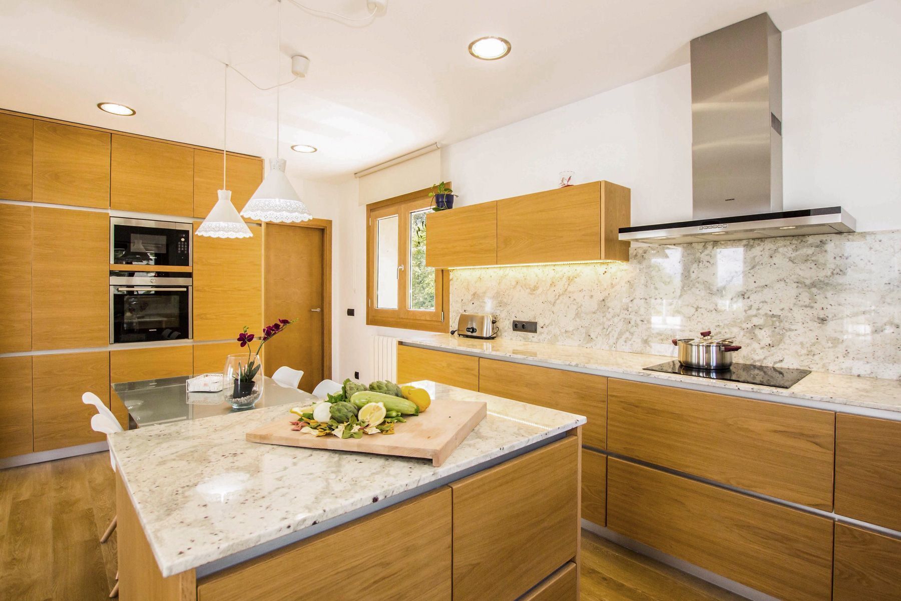 Una cocina moderna con armarios de madera, encimera de mármol, horno integrado y campana extractora de acero inoxidable.