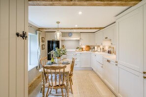 Een keuken met witte kasten, een houten eettafel met stoelen, een hanglamp en een raam met gordijnen.