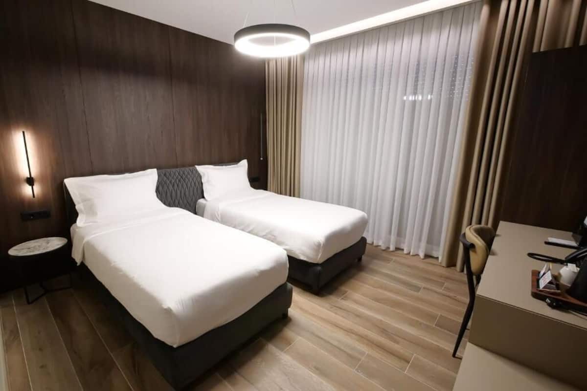 Ett hotellrum med två sängar, ett skrivbord, en stol och ett stort fönster med gardiner.