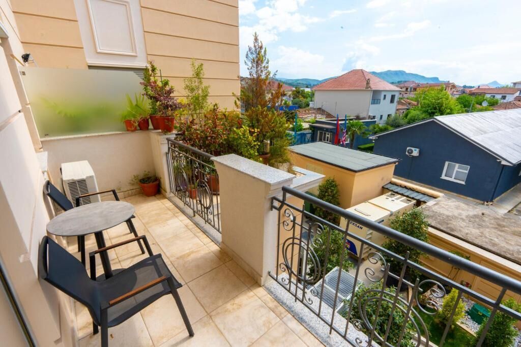 En balkon med et bord, stole, plantekrukker og udsigt til omkringliggende bygninger.