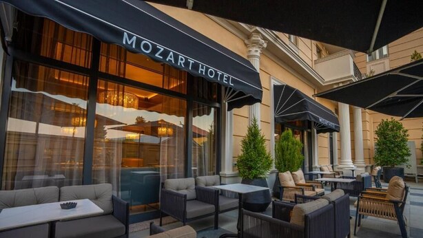 Uma área externa com sofás e mesas, um edifício com a placa 'MOZART HOTEL' e colunas.