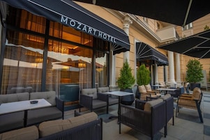 An outdoor seating area with sofas and tables, a building with a 'MOZART HOTEL' sign, and columns.