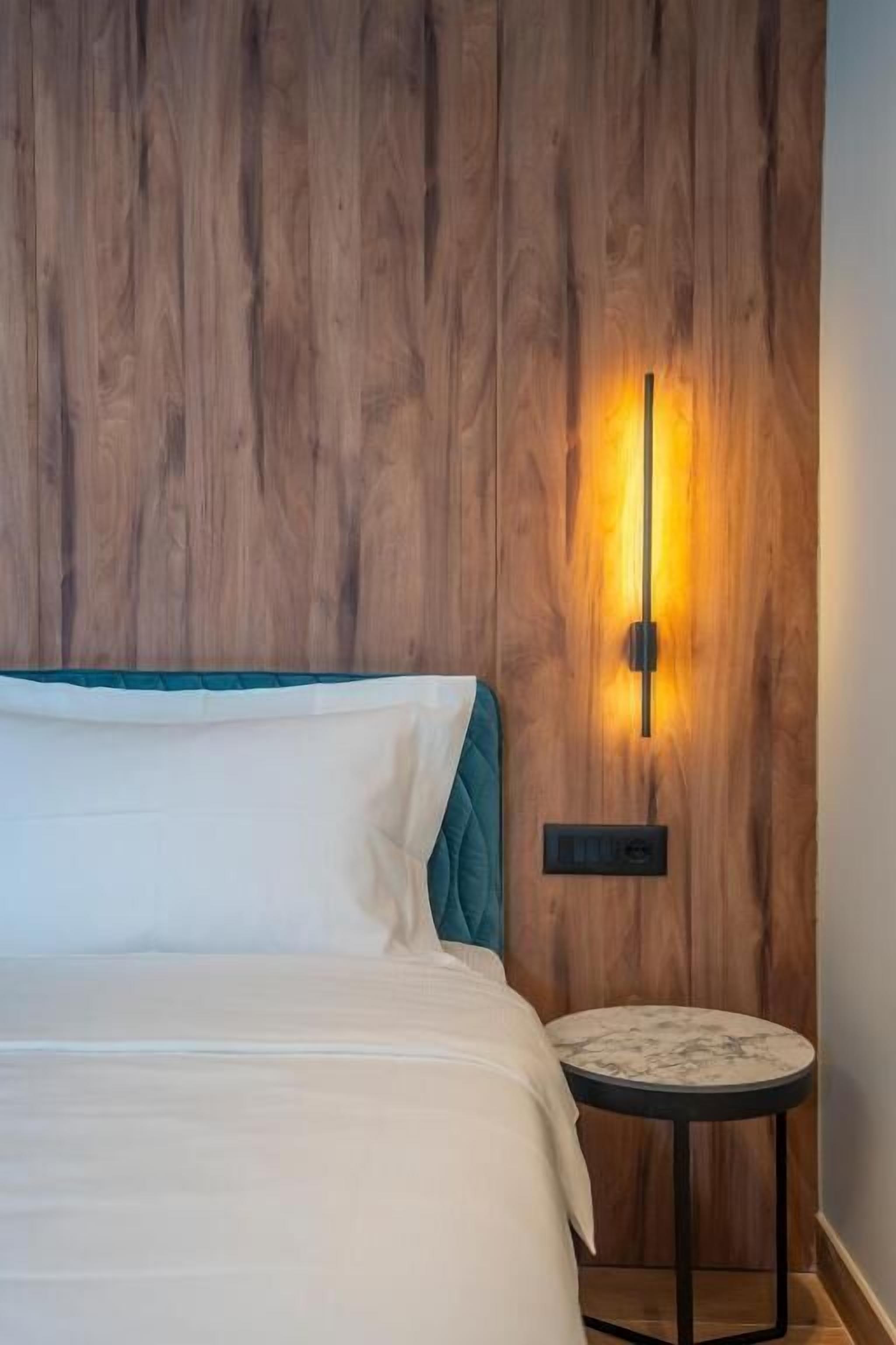 Uma cama com cabeceira azul-esverdeada, uma mesa redonda com tampo de mármore e um candeeiro fixo na parede.