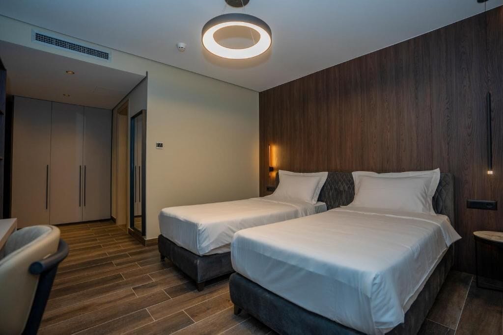 Ein modernes Hotelzimmer mit zwei Betten, einer Wand mit Holzvertäfelung, einer runden Deckenleuchte und einer sichtbaren Tür zu einem anderen Raum.