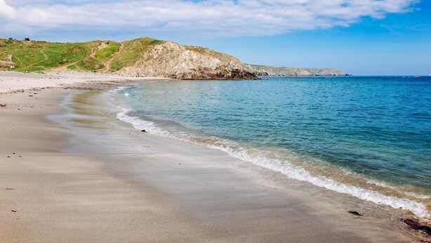 Una playa con agua cristalina, una orilla arenosa y una colina rocosa al fondo.