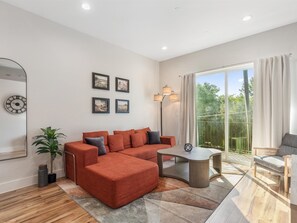A modern living room with a sectional sofa, a glass coffee table, and a large window with a view of greenery. - 1BR Getaway at Heart of Oaklawn EV Charger (Dallas)