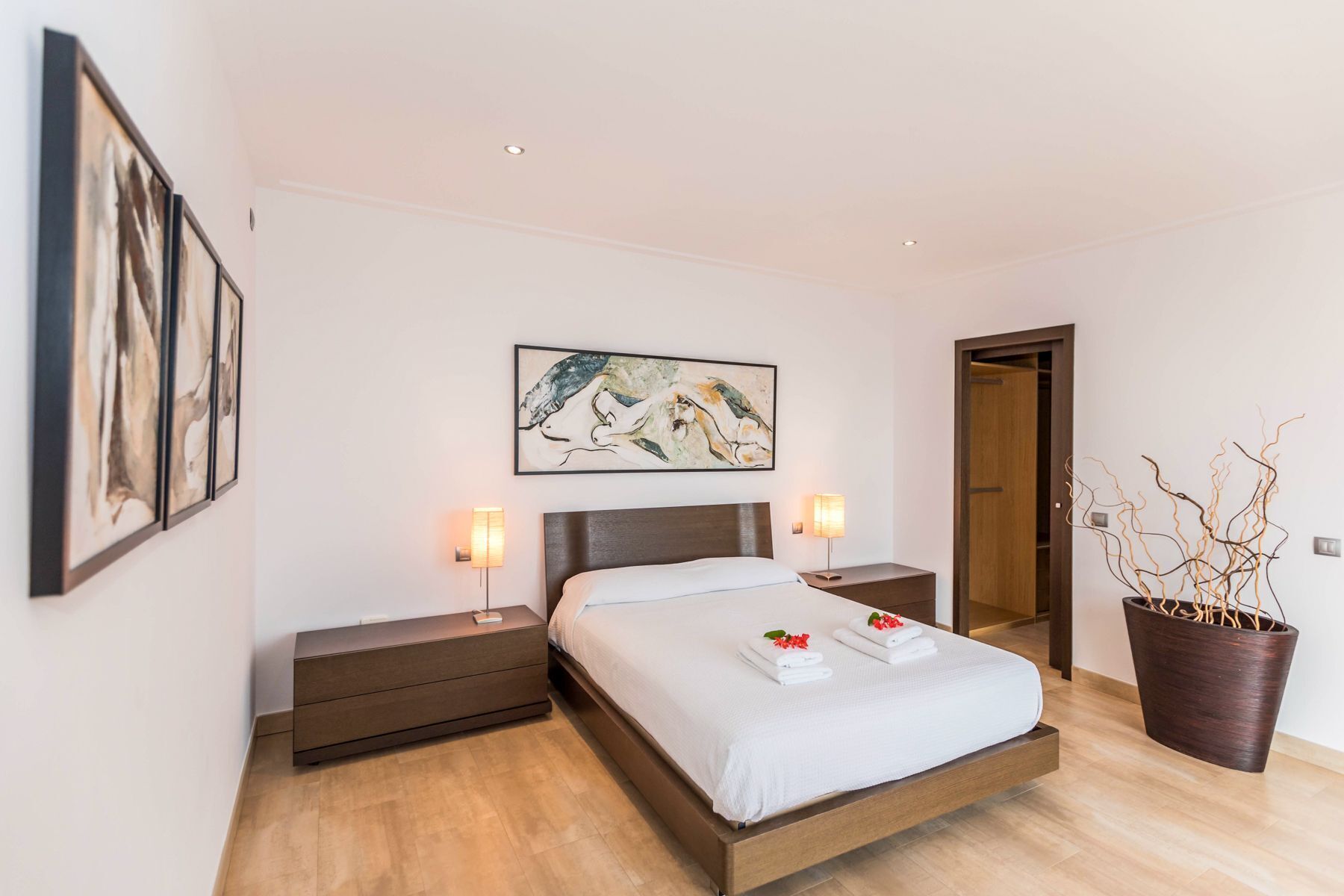 A modern bedroom with a bed, bedside tables, a nightstand, a potted plant, and artwork on the walls.
